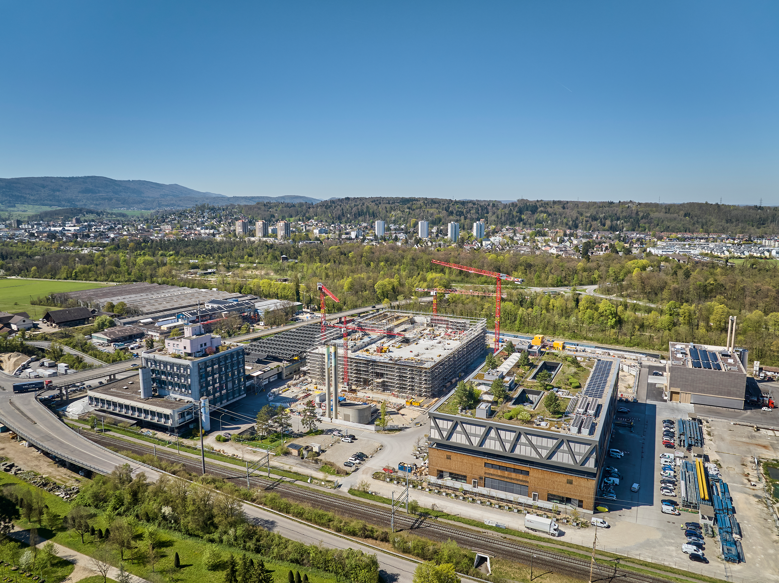 Aerial view of the uptownBasel site in Arlesheim in spring 2026 – Building 1 completed, Building 2 under construction, with a railway line and motorway link